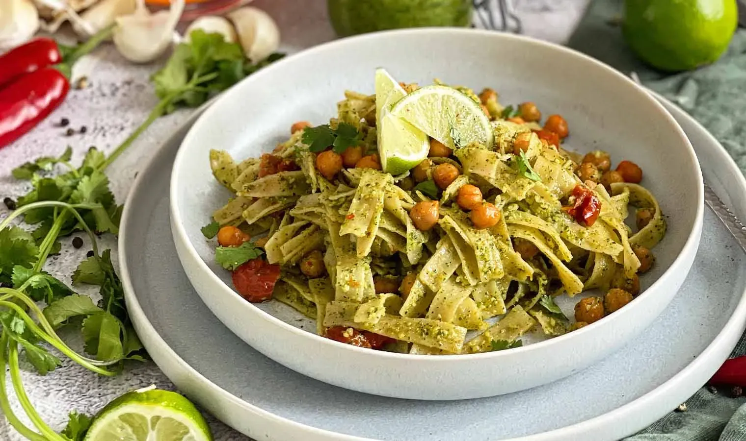 Bandnudeln mit Koriander-Minz-Pesto, Kichererbsen, Limettenscheiben und Kräutern auf einem Teller, daneben Zutaten