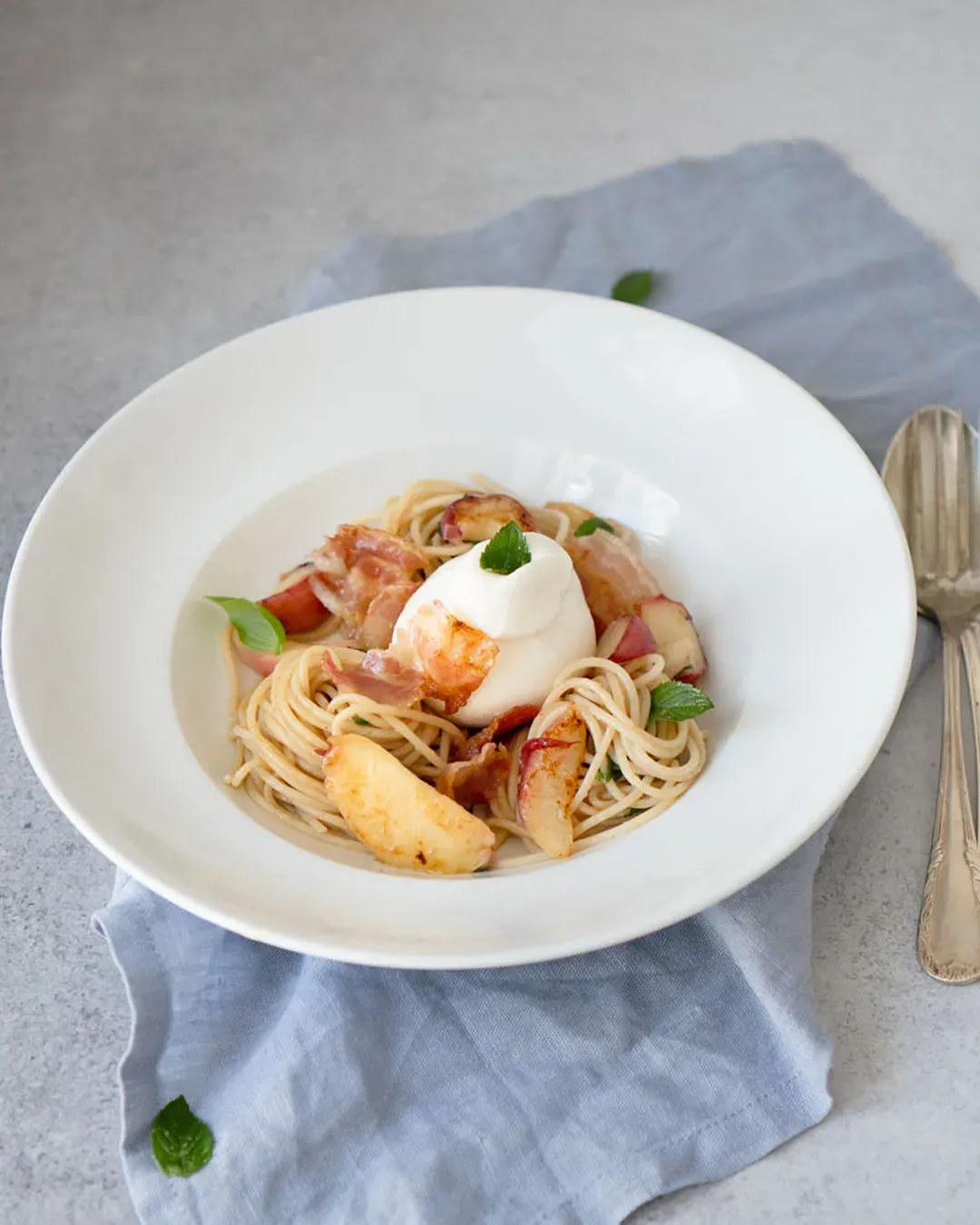 Dinkelspaghetti mit Pfirsichen, Pancetta und Burrata, mit einem Klecks Frischkäse auf blauem Tuch