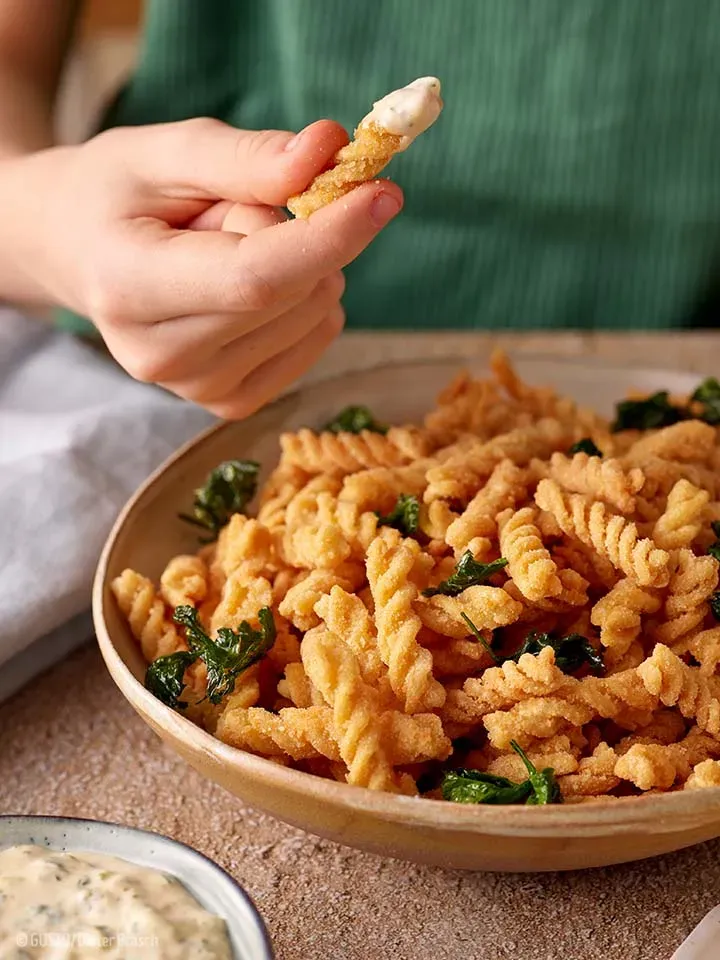 Hand hält einen mit Sauce tartare bedeckten, frittierten Dralli-Snack über einer Schüssel