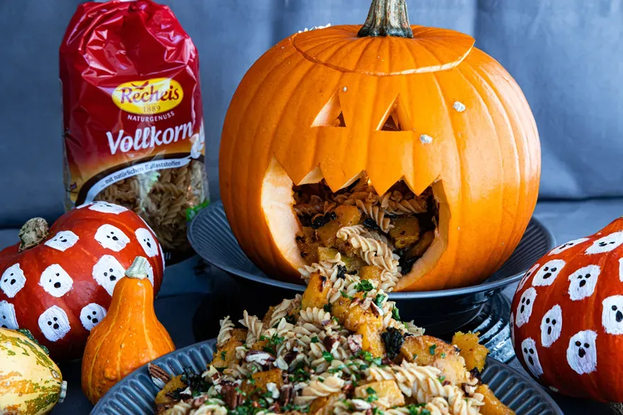 Halloween-Kürbis mit Gesicht, aus dem Vollkornnudeln mit Gemüse quellen, umgeben von bemalten Kürbissen