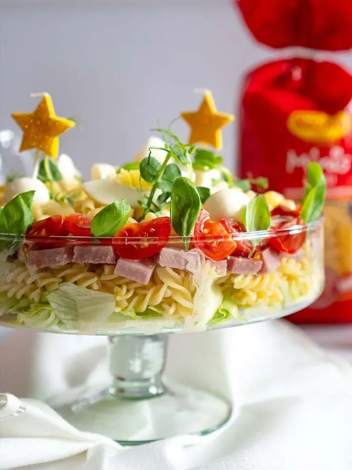 Geschichtete Nudelsalattorte mit Tomaten, Schinken und grünen Blättern in einer Glasschale