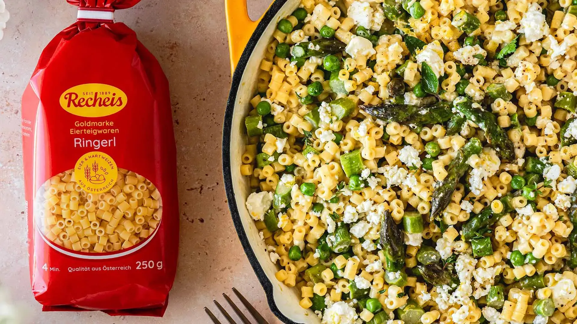 Aufsicht auf die One-Pot Spargel-Nudel-Pfanne mit Feta neben einer Packung Recheis Goldmarke Ringerl