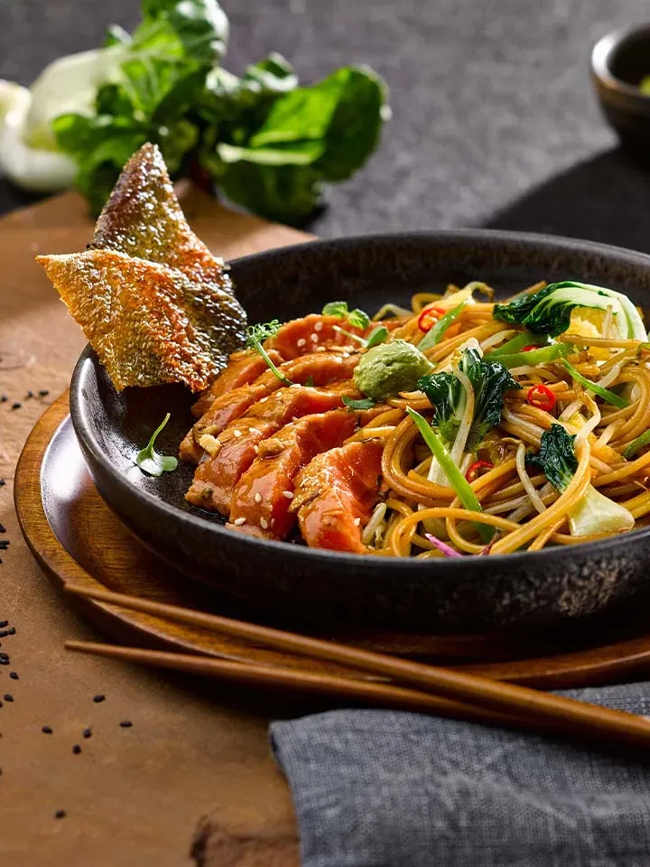 Asia-Nudelgericht mit Lachs, Gemüse und knuspriger Fischhaut in schwarzer Schale