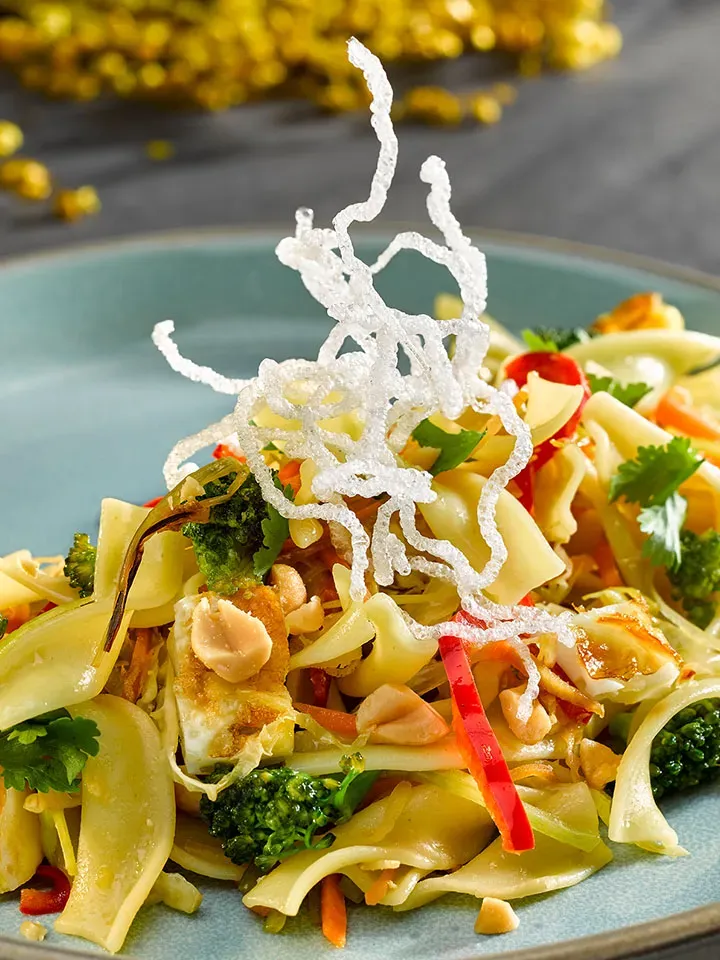 Asia-Nudelpfanne mit Gemüse, Erdnüssen und knusprigen Reisnudeln auf einem blauen Teller, Gastrorezept