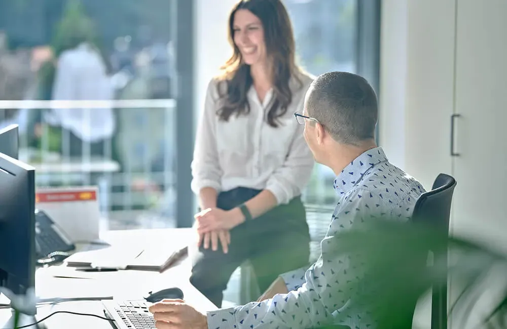 Zwei Kollegen unterhalten sich freundlich im hellen Büro mit Computerarbeitsplatz