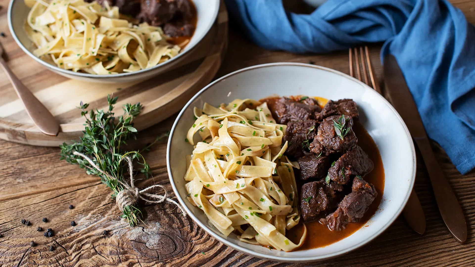 Bandnudeln mit Gulasch, angerichtet auf einem Teller auf Holztisch mit Kräutern