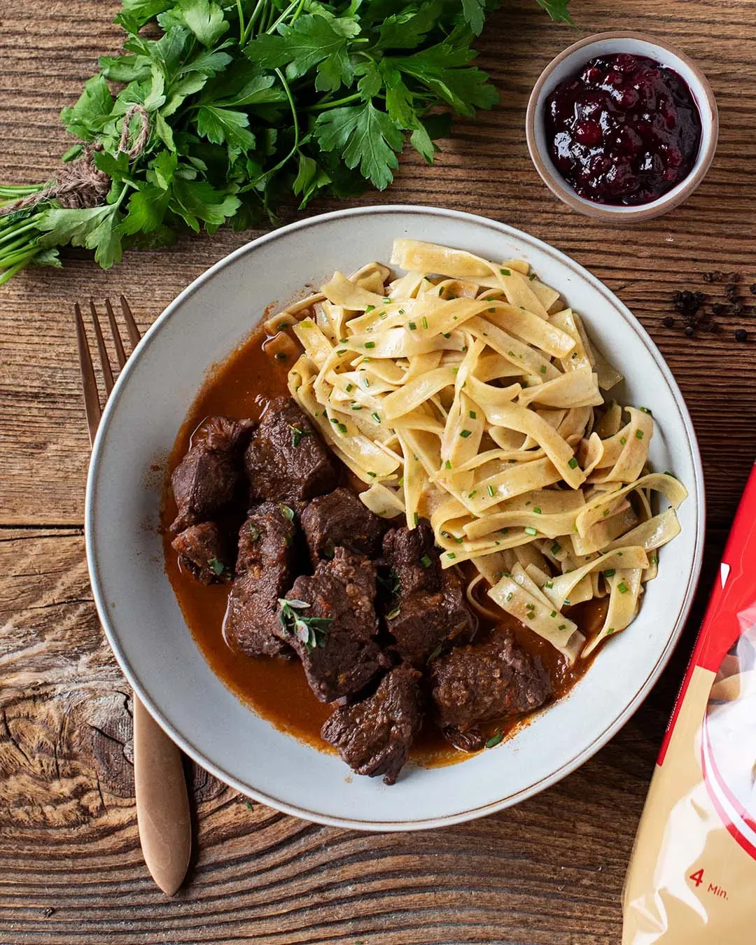 Bandnudeln mit Rindergulasch und Preiselbeersoße auf einem Teller auf Holztisch