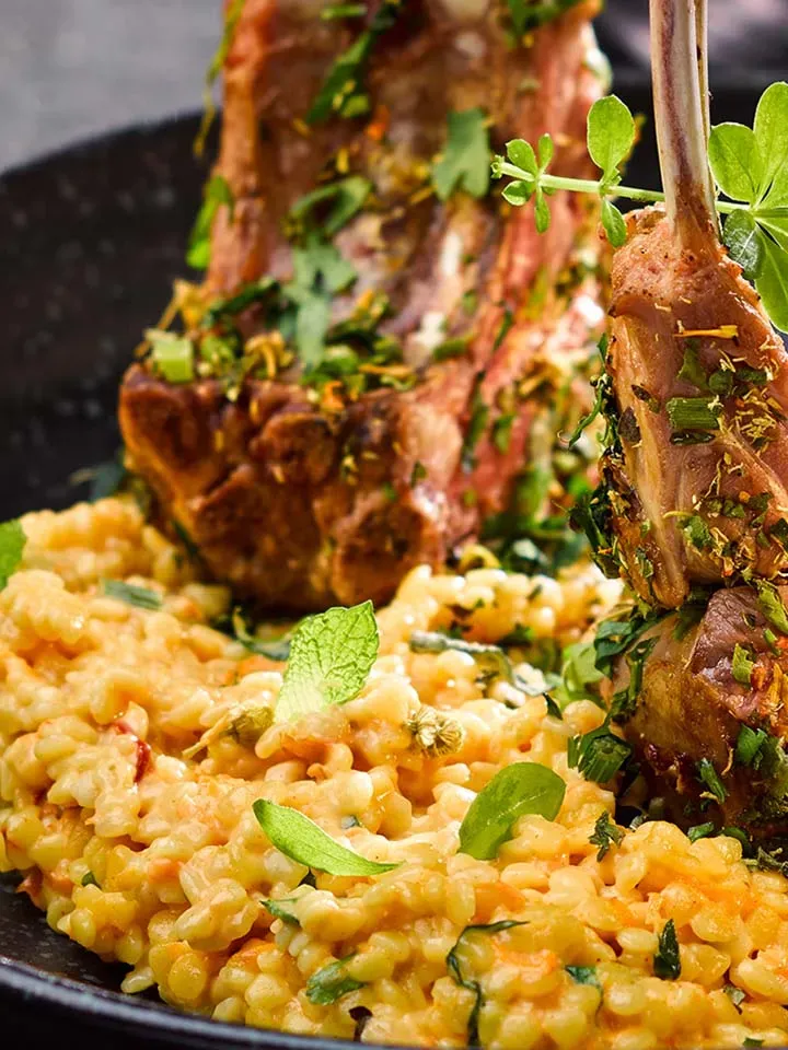 Nahaufnahme: Berglamm-Koteletts im Kräutermantel mit cremigem Risotto auf einem schwarzen Teller