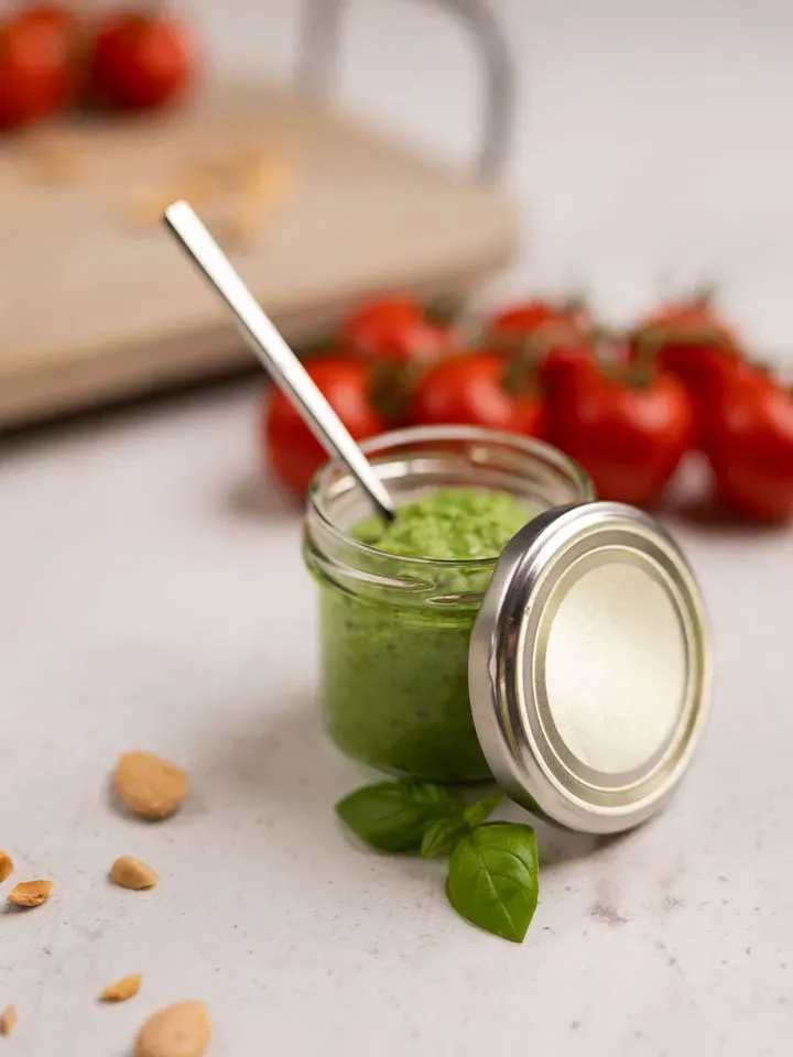 Selbstgemachtes Brokkoli-Pesto im Glas mit Löffel, Basilikum und Tomaten im Hintergrund