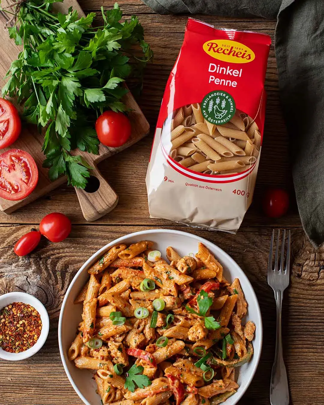 Cajun Chicken mit Dinkel-Penne, frischen Tomaten, Kräutern und Gewürzen auf einem Teller auf Holztisch