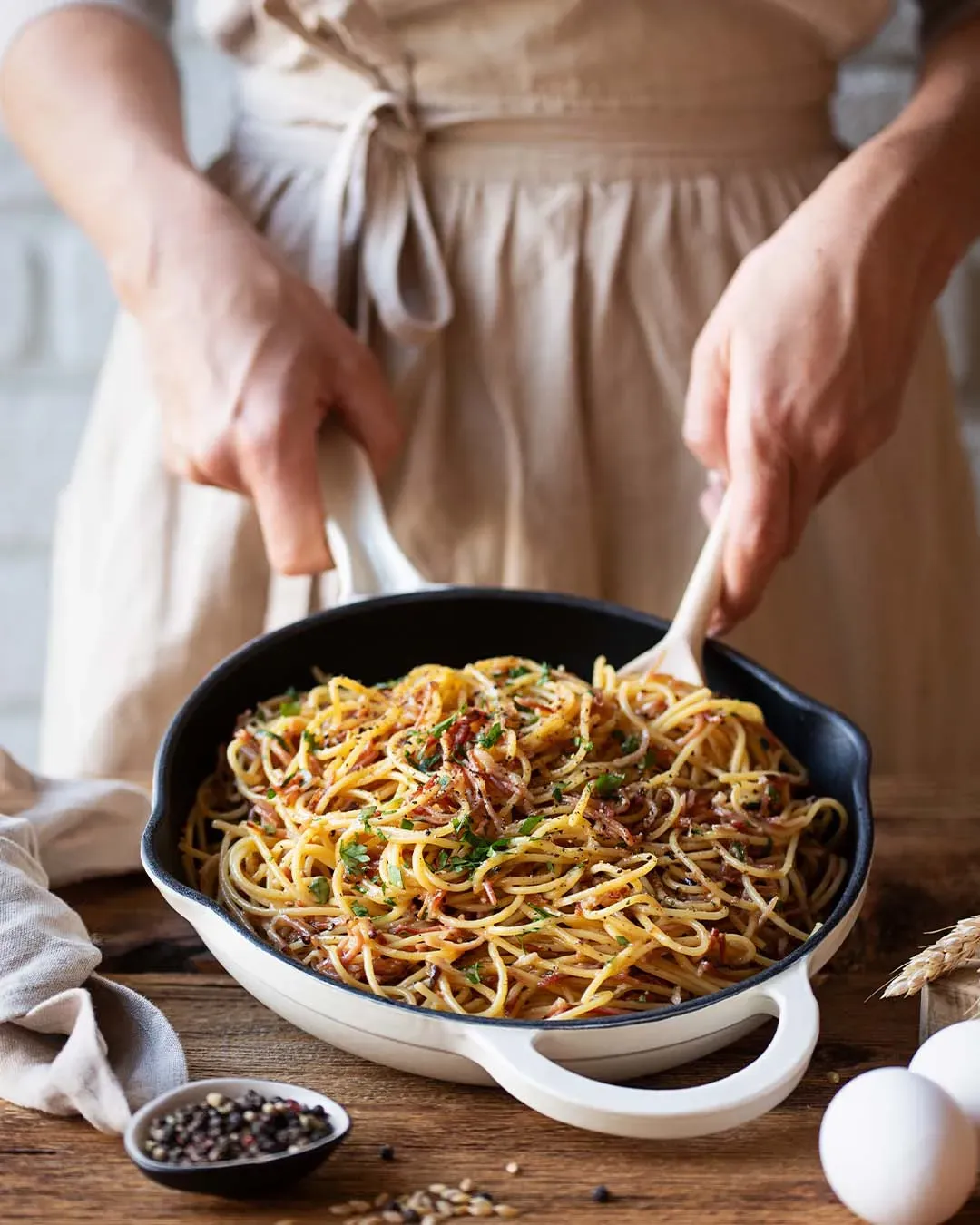 Person hält eine Pfanne mit Carbonara aus Tirol, Spaghetti mit Speck, auf Holztisch mit Eiern und Pfeffer