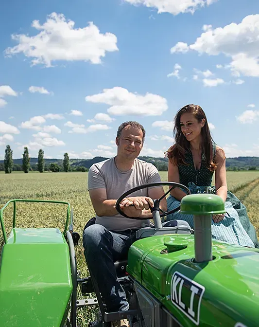 Johannes Pock am Steuer eines grünen Oldtimer-Traktors, neben ihm sitzt Cooking Catrin, im Hintergrund ein Feld