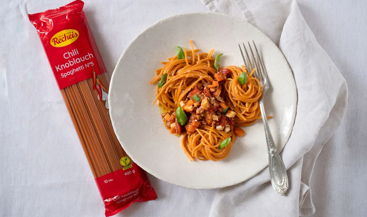 Vegane Chili-Knoblauch-Spaghetti Bolognese mit Sonnenblumenhack, daneben die Packung auf weißem Tuch