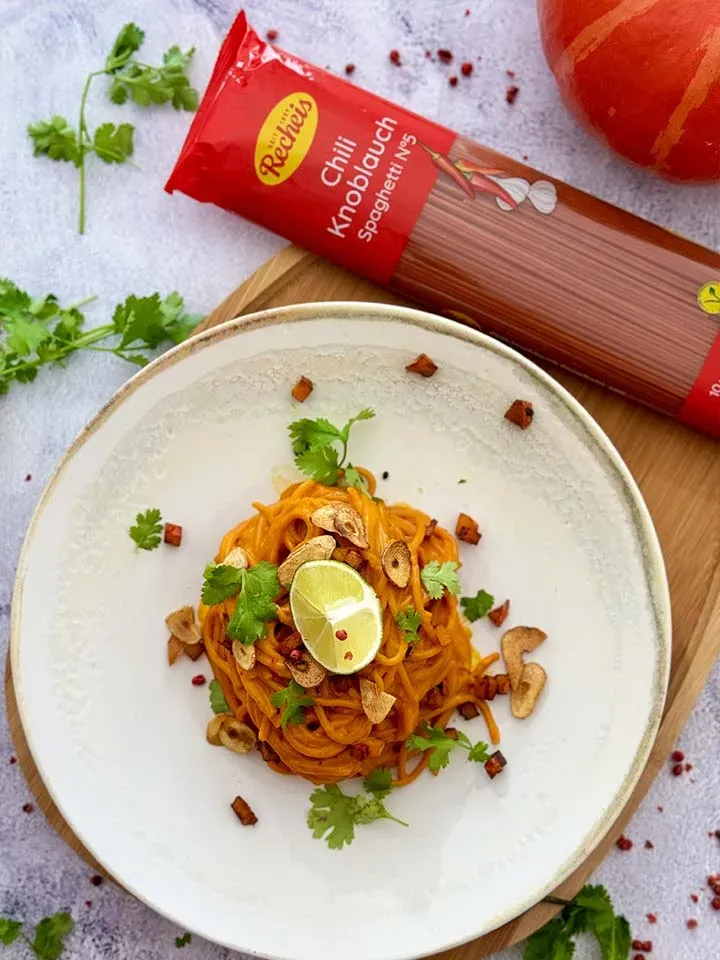 Chili-Knoblauch-Spaghetti mit Kürbis und Gochujang, mit Limette und Kräutern garniert, neben einer Nudelpackung