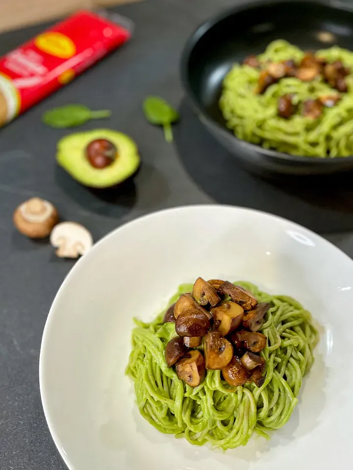 Cremige Avocado-Spaghetti mit gebratenen Champignons und Urkorn-Emmer-Nudeln auf weißem Teller, Zutaten im Hintergrund
