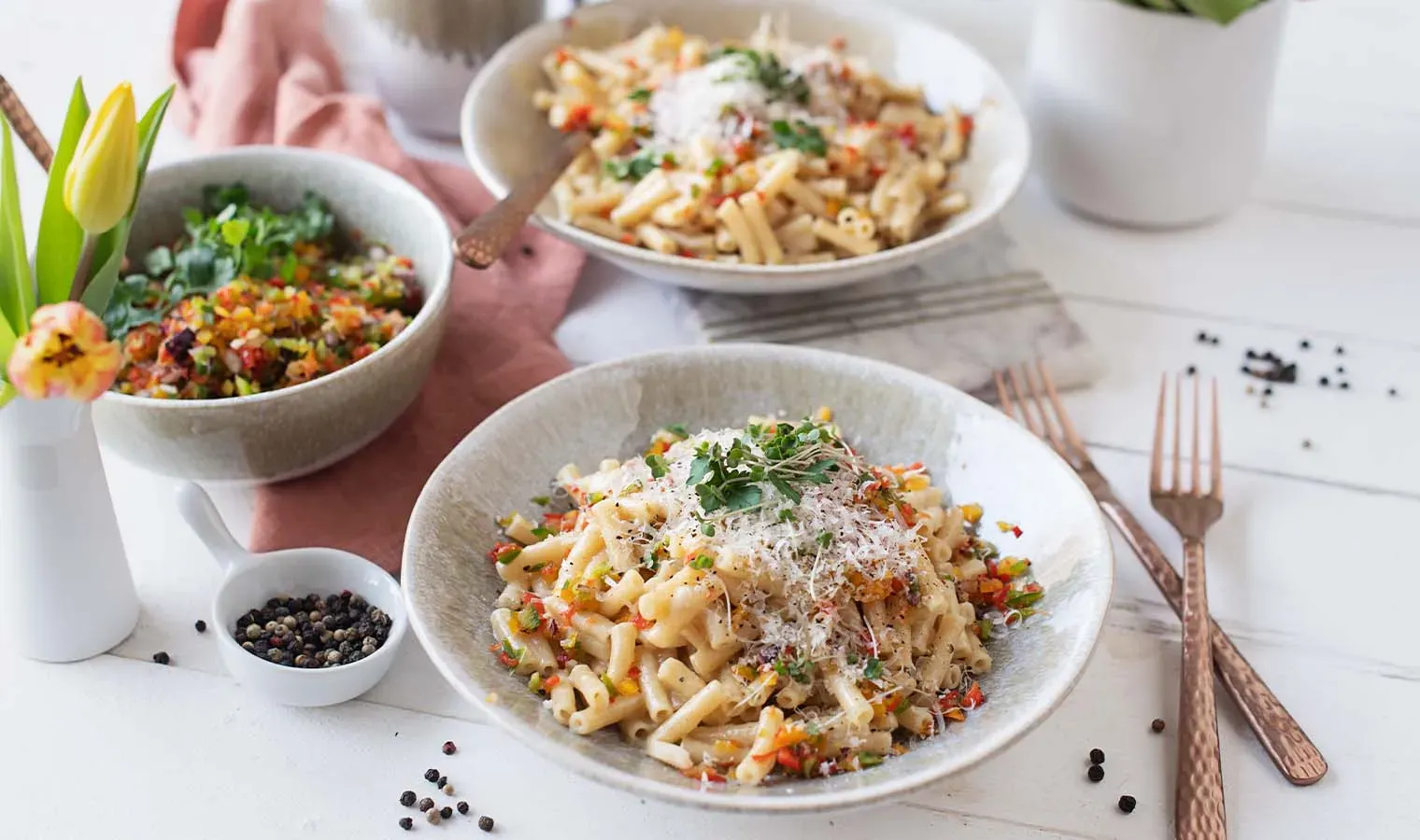Dinkel-Makkaroni Cacio e Pepe mit Gemüse auf zwei Tellern auf weißem Tisch mit Besteck und Tulpen