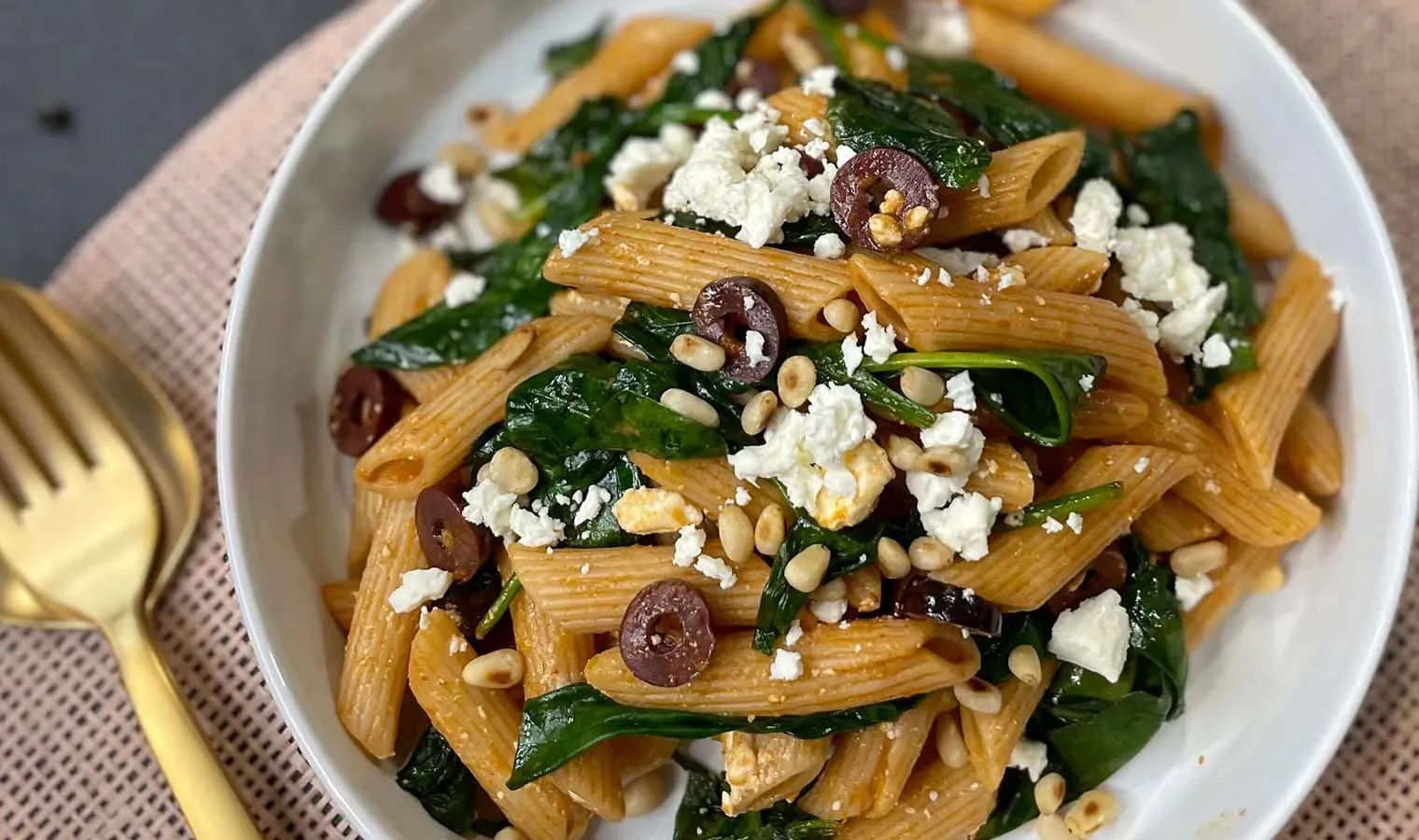 Dinkel-Penne mit Harissa, Spinat, Oliven, Feta und Pinienkernen auf einem Teller