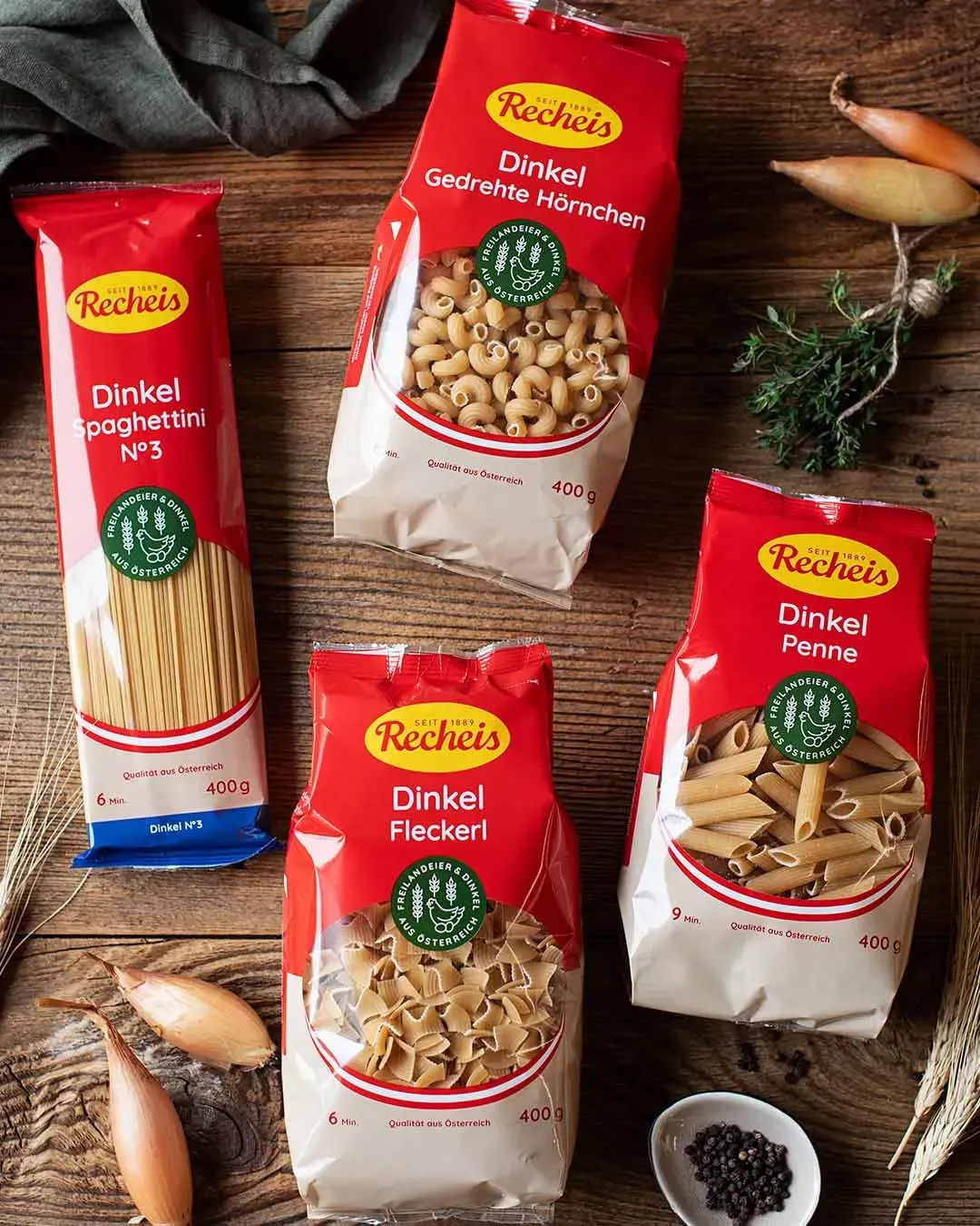 Verschiedene Packungen Recheis Dinkelnudeln auf einem Holztisch mit Zwiebeln und Kräutern, von oben