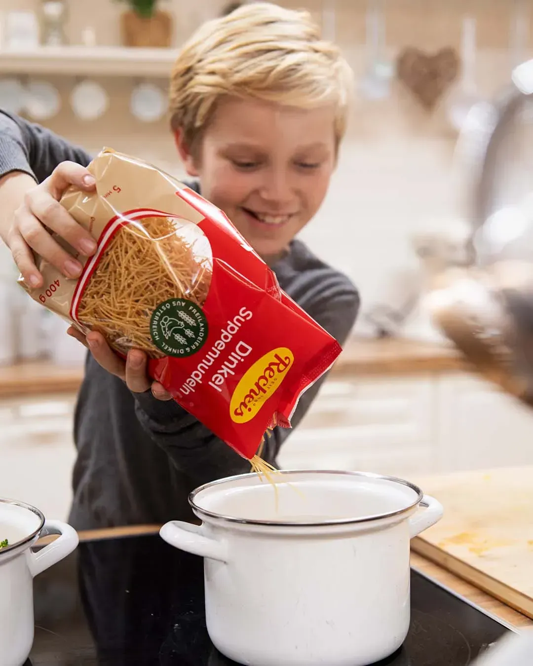 Ein Bub schüttet Recheis Dinkel Suppennudeln aus der Packung in einen Kochtopf auf dem Herd