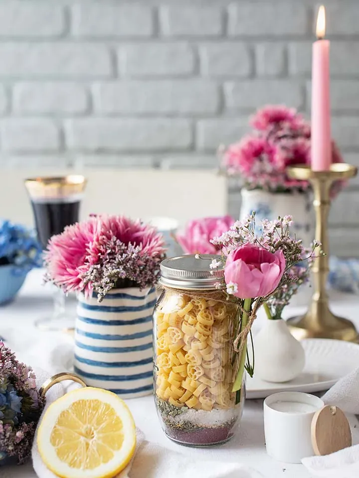 Nudeln im Glas zum Verschenken auf einem Tisch mit Blumen, Kerze und Zitronenhälfte auf weißem Tuch