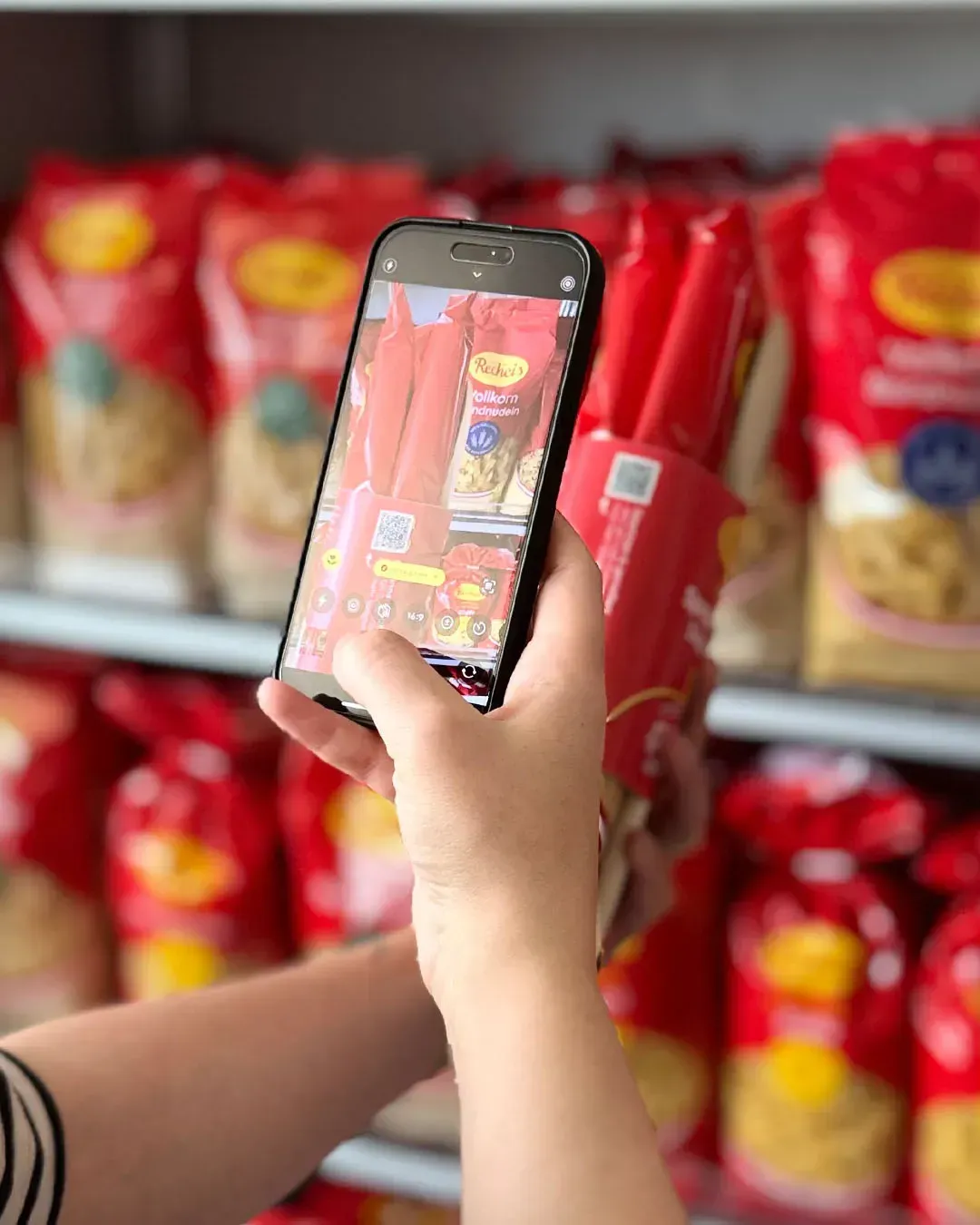 Hand scannt mit dem Smartphone den QR-Code auf einer Recheis Spaghetti-Doppelpackung im Nudelregal