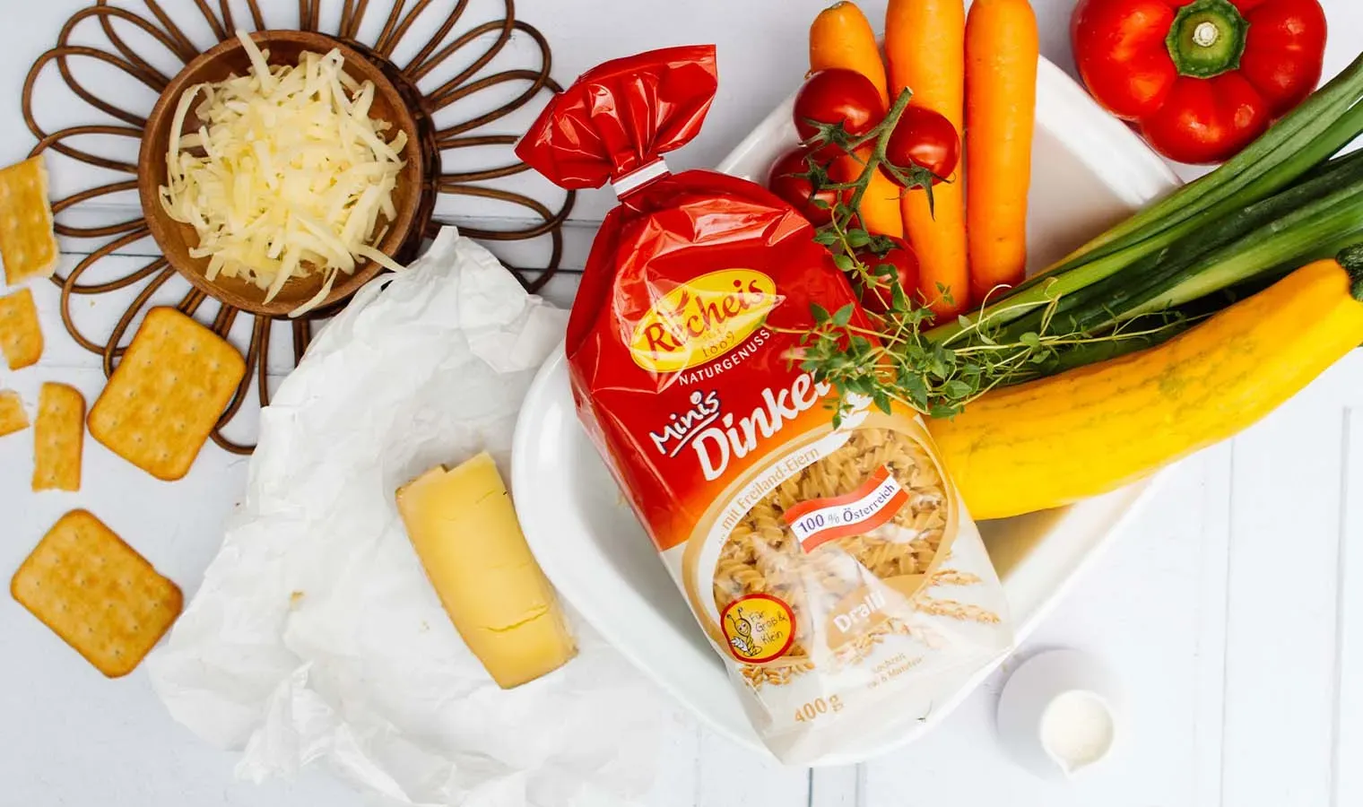 Dralli-Nudelauflauf mit Zutaten wie Nudeln, Käse, Gemüse und Crackern auf weißem Untergrund