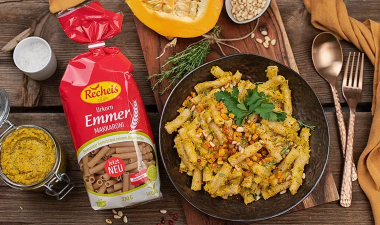 Emmer-Makkaroni mit Kürbis auf Teller, daneben Packung und Gewürze auf Holzuntergrund