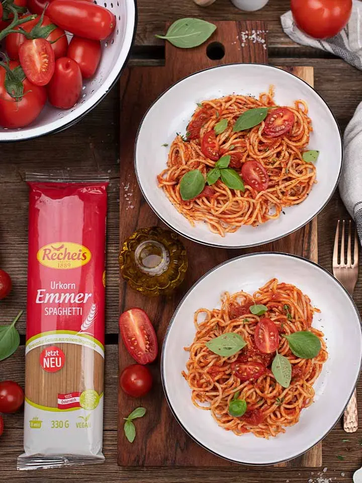 Emmer-Spaghetti Napoli auf zwei Tellern, daneben rohe Tomaten und eine Spaghettipackung