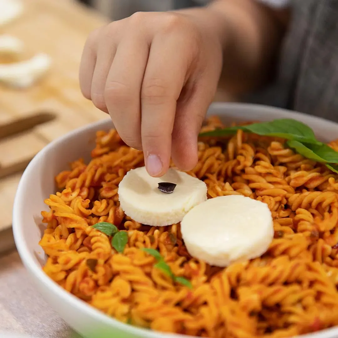 Hand legt ein Olivenstück auf Mozzarella in einer Eulen-Bowl mit Nudeln