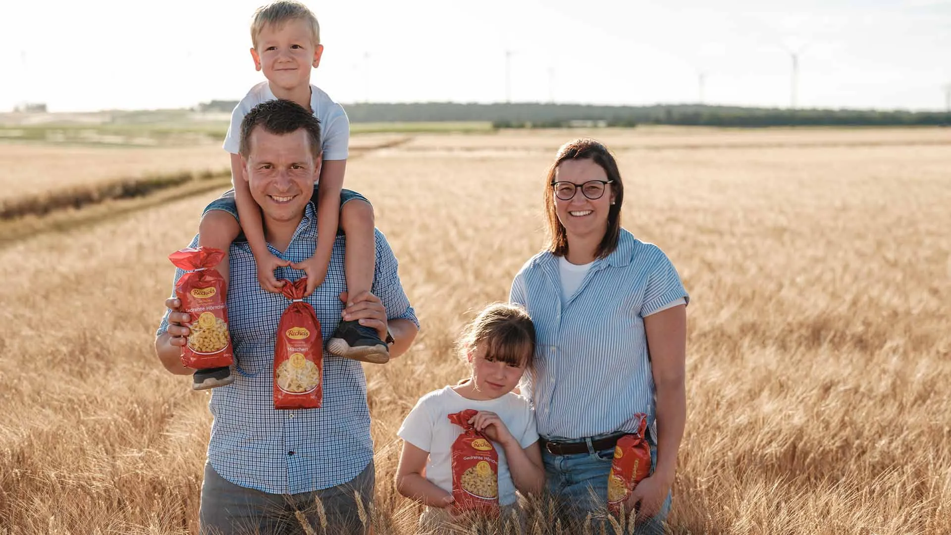Familie mit Kindern hält Nudeltüten auf einem Feld mit Windrädern im Hintergrund