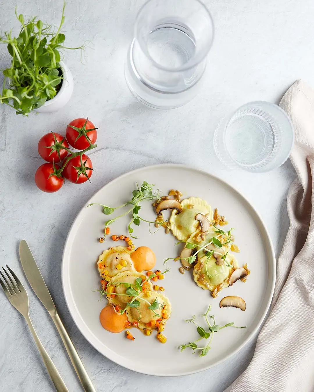 Fiori zweierlei als Vorspeise: Teller mit Ravioli, Pilzen und Kräutern neben Tomaten und Wasserglas