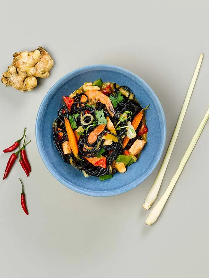 Fischcurry-Pastabowl: blaue Schüssel mit schwarzer Pasta, Garnelen und Gemüse, daneben frische Zutaten