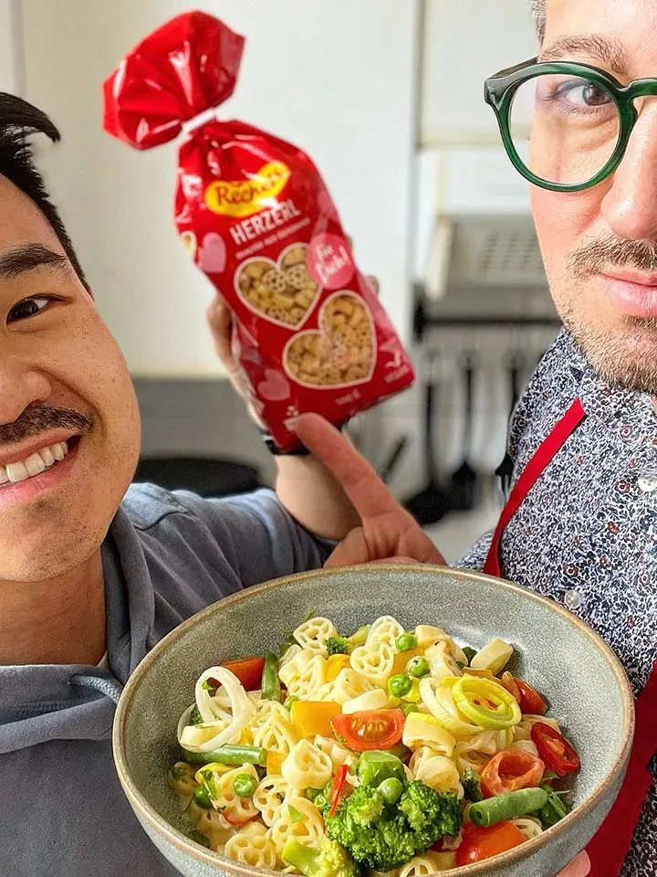 Zwei Männer zeigen Herznudeln und eine Schüssel mit Frühlingsgemüse und Pasta