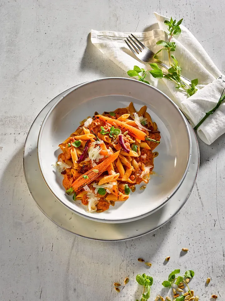 Alpenglühen-Penne mit Karotten und Kräutern auf hellem Untergrund mit Besteck