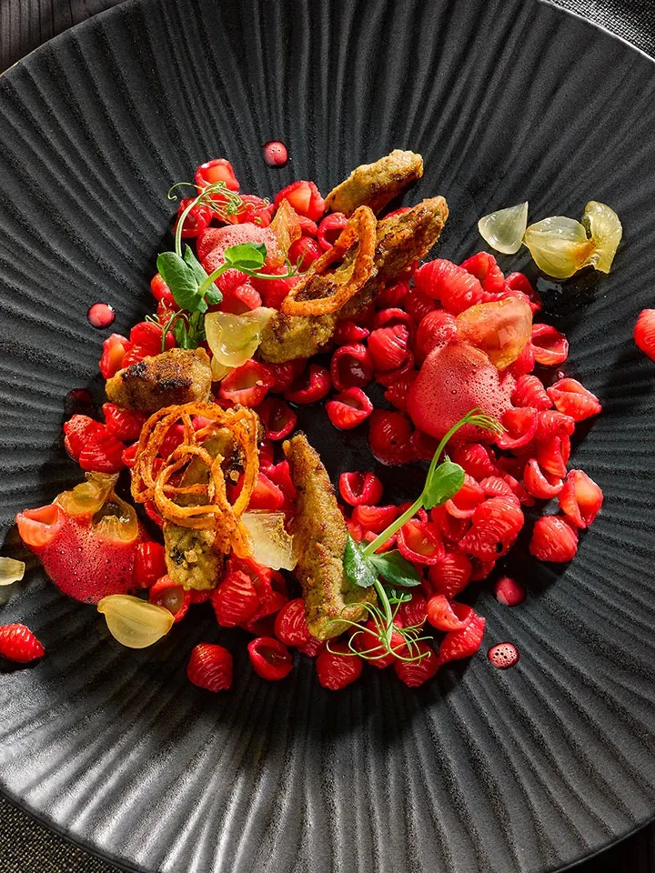 Kalbsleber mit Roter Beete und roten Muschelnudeln, mit frittierten Stücken und Kräutern auf schwarzem Teller
