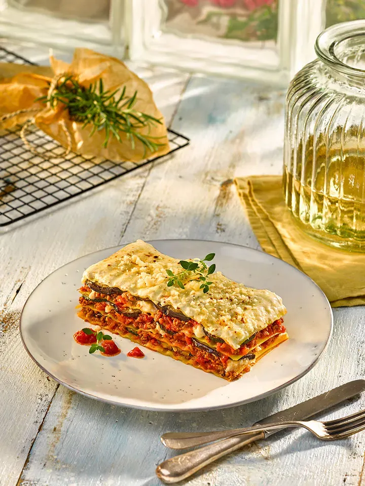 Stück Lasagne al forno mit frischen Kräutern auf einem Teller auf Holztisch, Gastrorezept