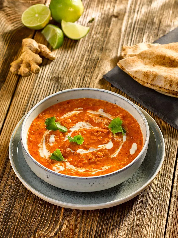 Rotes Linsen-Dal: Schüssel mit Linsensuppe, daneben Limetten, Ingwer und Fladenbrot auf Holztisch