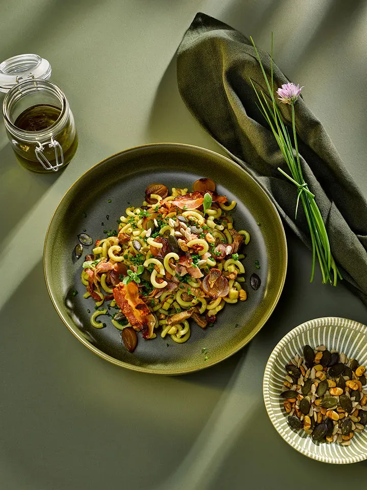 Steirischer Nudelsalat mit Speck und Kräutern, daneben ein Glas Öl und eine Schale Samen