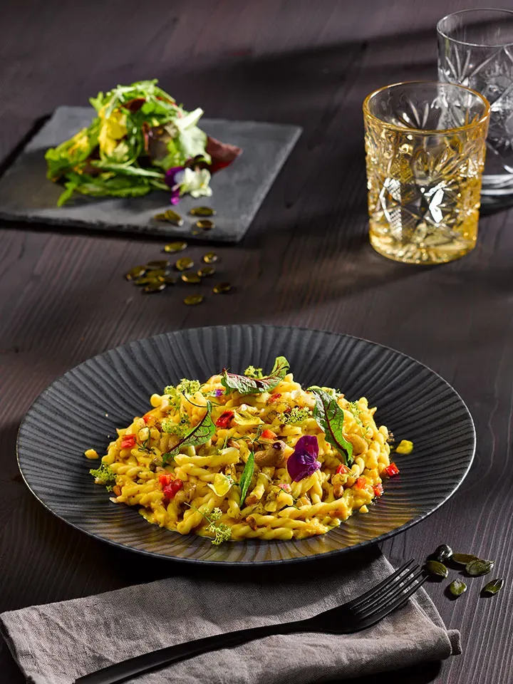 Treccine in Kürbis-Grillen-Sahne-Sauce mit Kräutern, dazu Salat auf Steinplatte und zwei Gläser auf dunklem Tisch