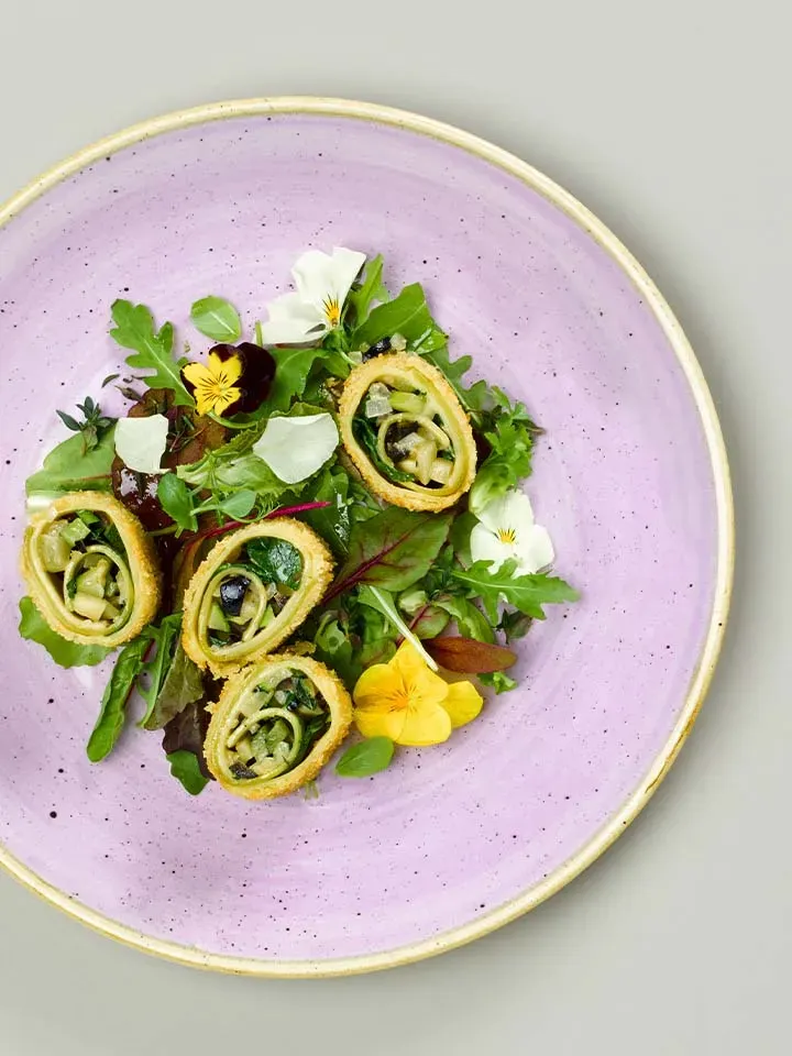Gebackene Lasagneschnecken mit Zucchini, panierte Rollen mit Salat und essbaren Blüten auf lila Teller