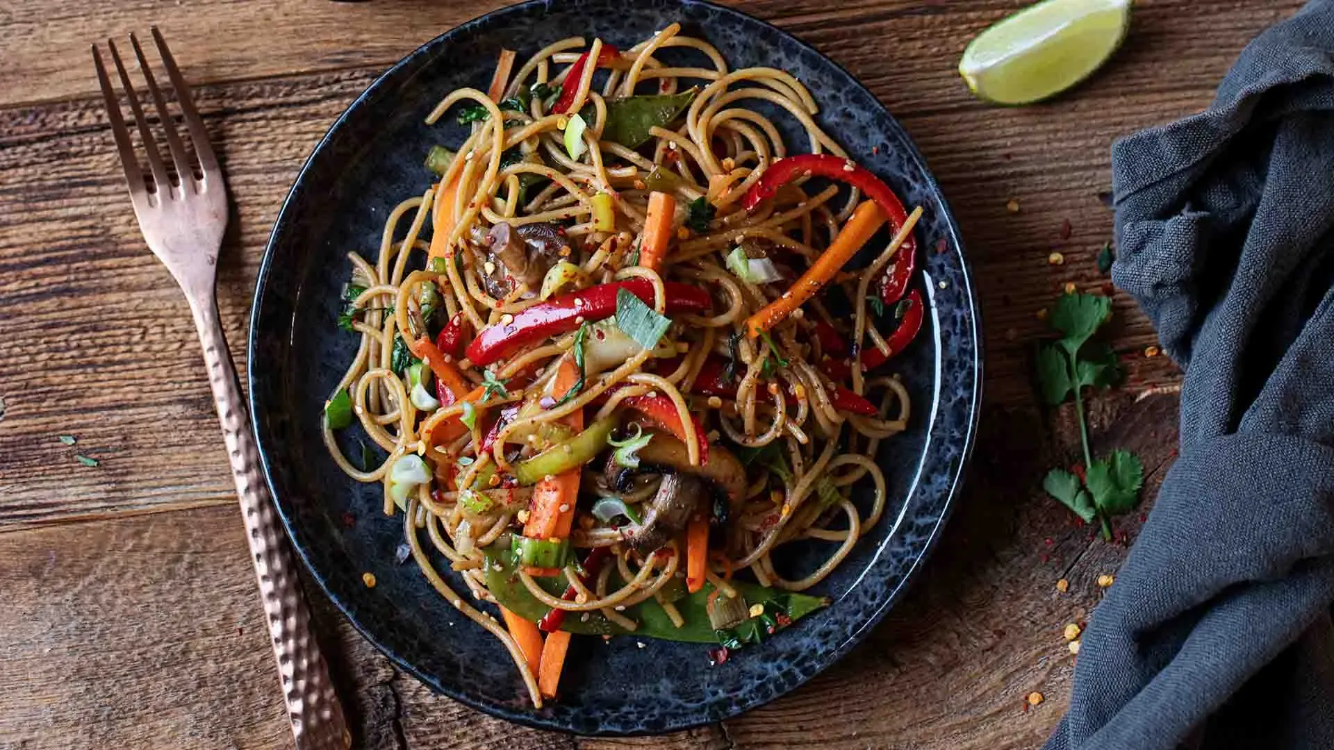 Gebratene Spaghetti mit würzigem Gemüse auf einem Teller auf Holztisch mit Gabel und Limettenspalte