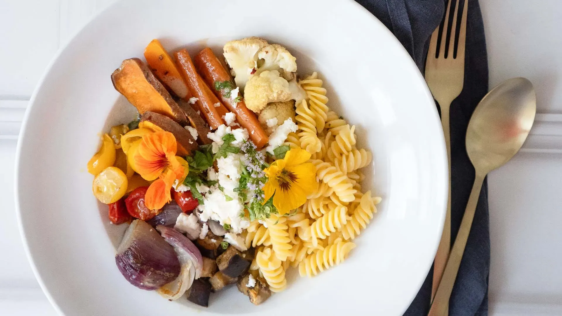 Gemüse-Nudelbowl mit geröstetem Gemüse und essbaren Blüten auf einem Teller neben Besteck