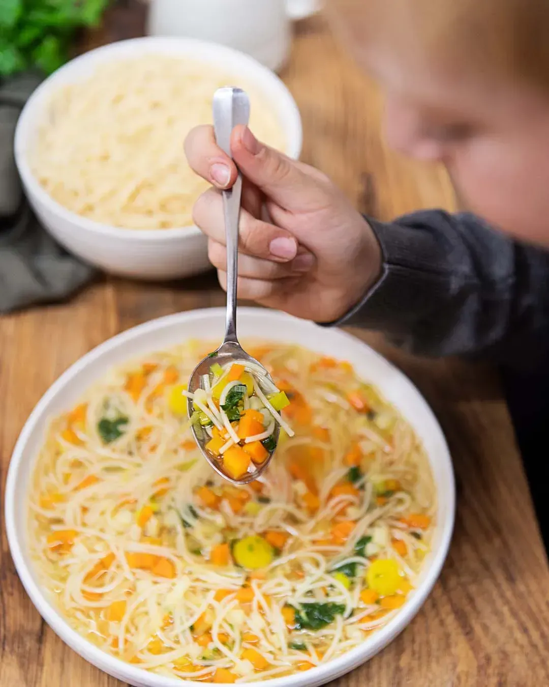 Ein Kind hält einen Löffel mit Suppennudeln und Gemüsewürfeln über einen Teller Gemüsesuppe