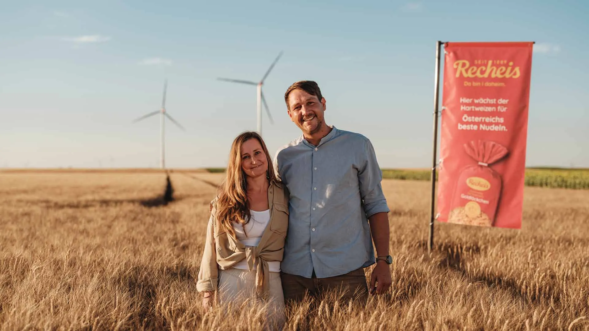 Paar steht in einem Weizenfeld mit Windrädern und Werbebanner für Nudeln im Hintergrund