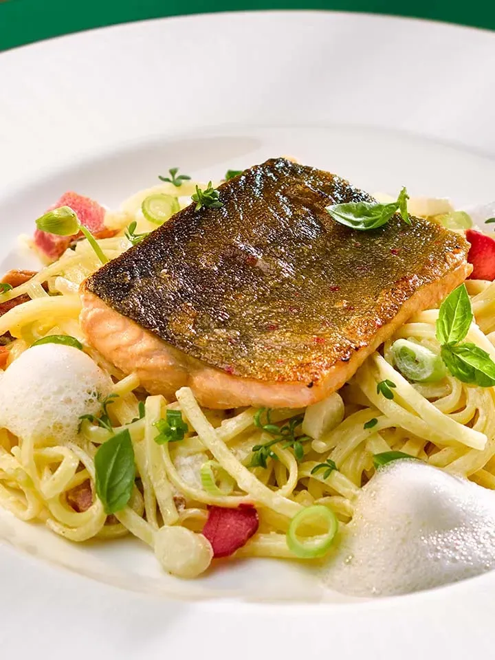 Nahaufnahme: geschmolzenes Saiblingsfilet mit Hausmacher-Nudeln, Kräutern und Schaum auf weißem Teller