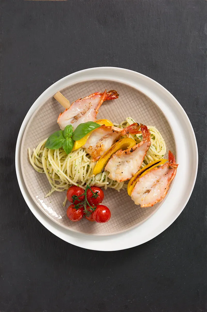 Glutenfreie Spaghetti mit Basilikumpesto und Scampispieß, Kirschtomaten und Basilikum auf weißem Teller