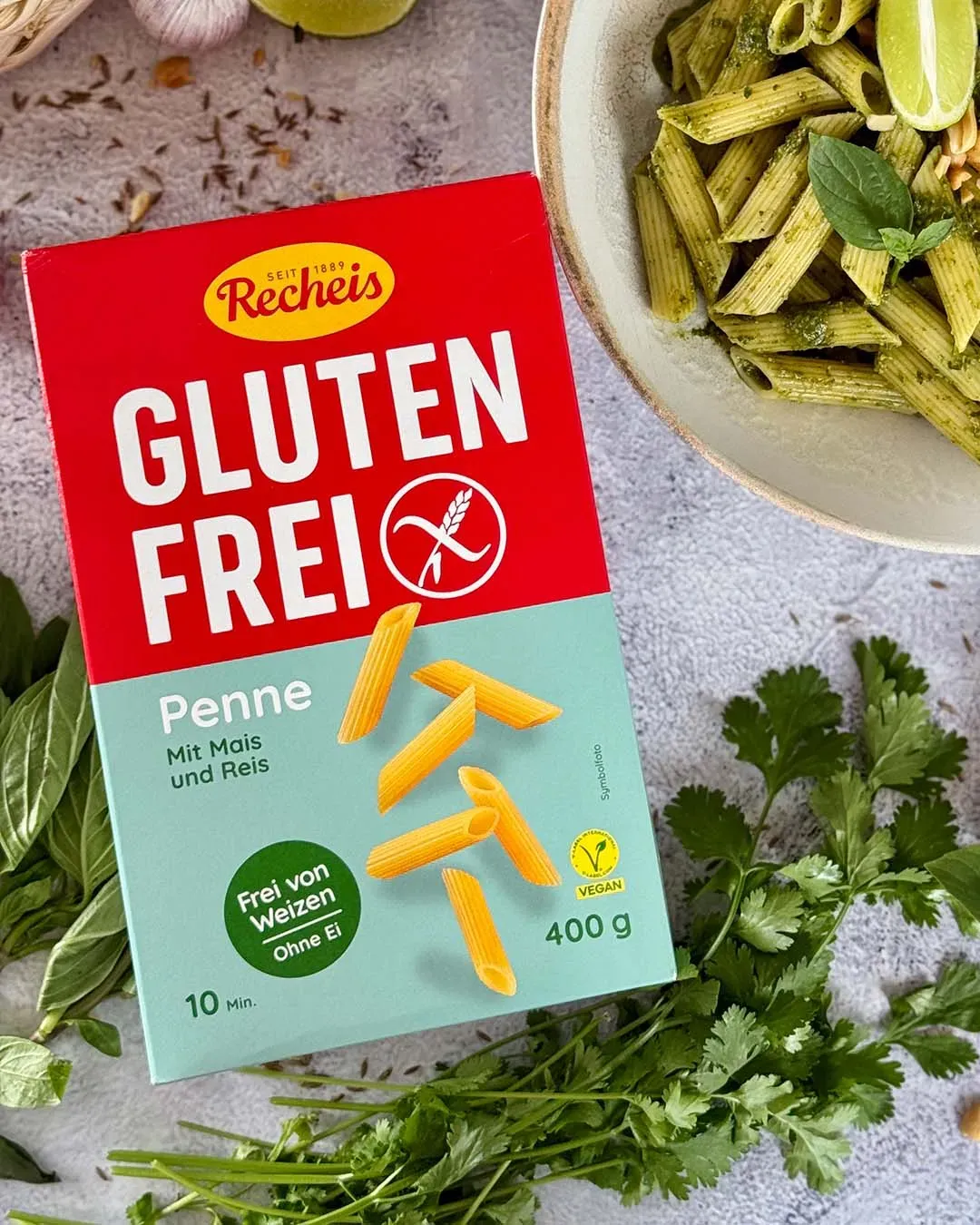 Packung glutenfreie Penne neben einem Teller mit Thai-Basilikum-Pesto-Pasta und frischen Kräutern