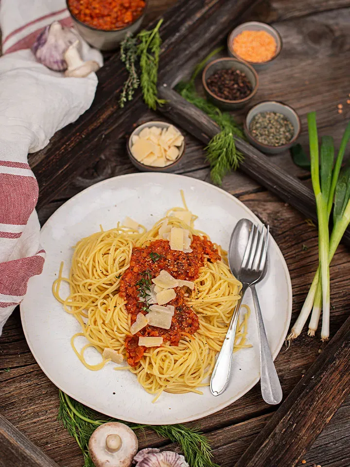 Glutenfreie Linsenbolognese mit Spaghetti und Parmesan auf einem Teller auf Holztisch, dazu Kräuter und Gewürze