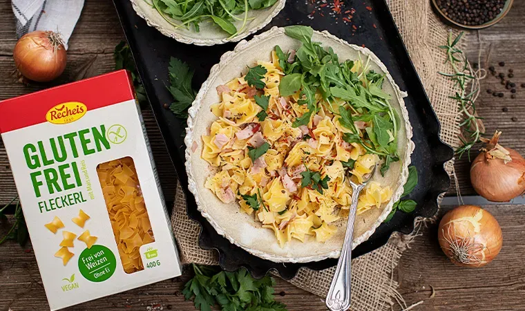 Glutenfreie Schinkenfleckerl mit frischem Rucola auf einem Teller neben Packung und Zwiebeln