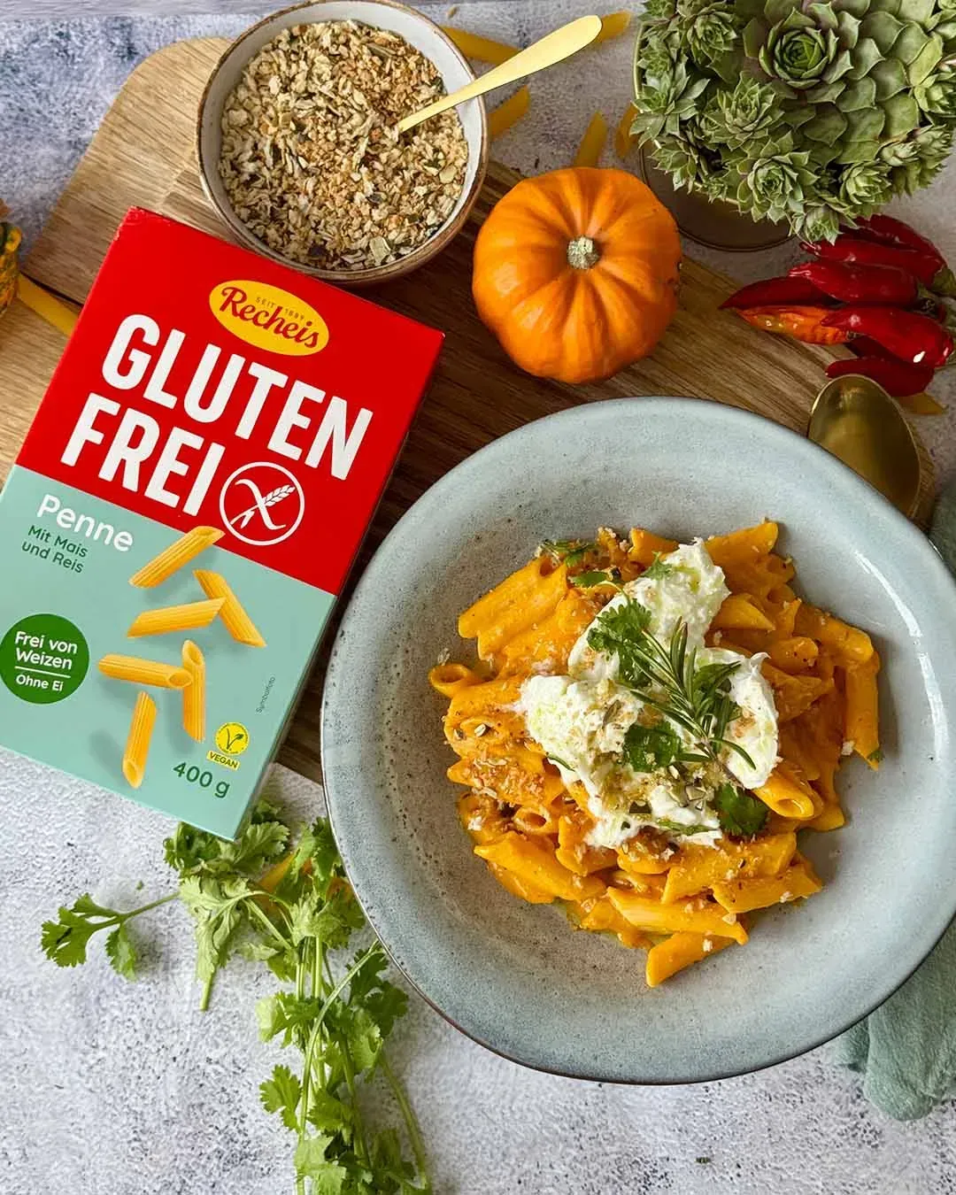 Aufsicht auf Kürbis-Ofenpasta mit Burrata, daneben eine Packung Recheis Glutenfrei Penne, ein Kürbis und Chilischoten